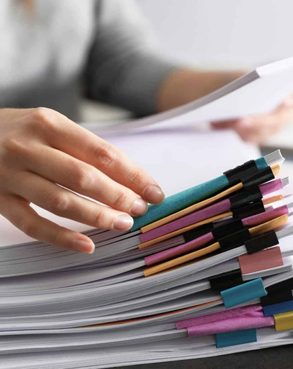 Person blättert in einem Stapel Unterlagen mit farbigen Registermarkierungen auf einem Schreibtisch.